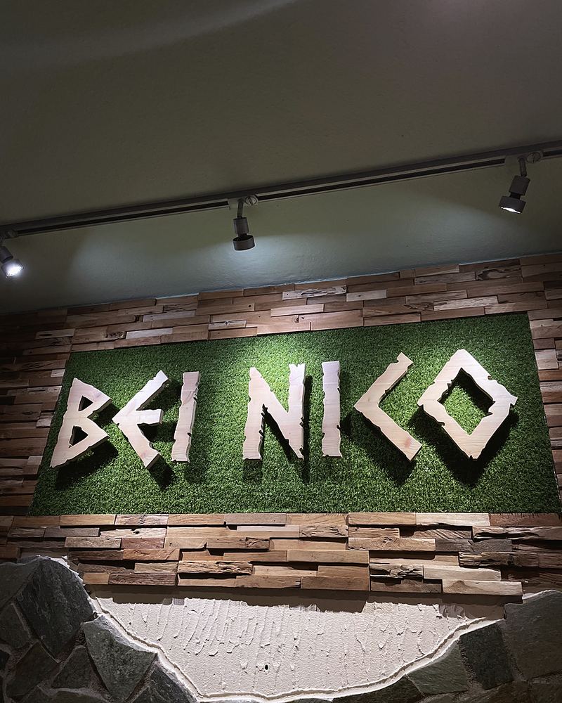 Der Schriftzug „Bei Nico“ aus hellem Holz auf grüner Wand neben Natursteinmauer